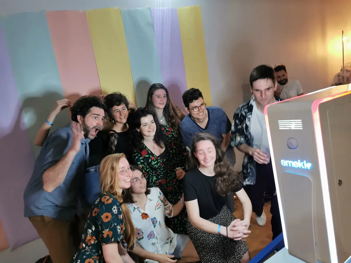 Une douzaine de personnes se serrent devant la borne Smakk pour une photo de groupe, mur de bandes pastel en fond