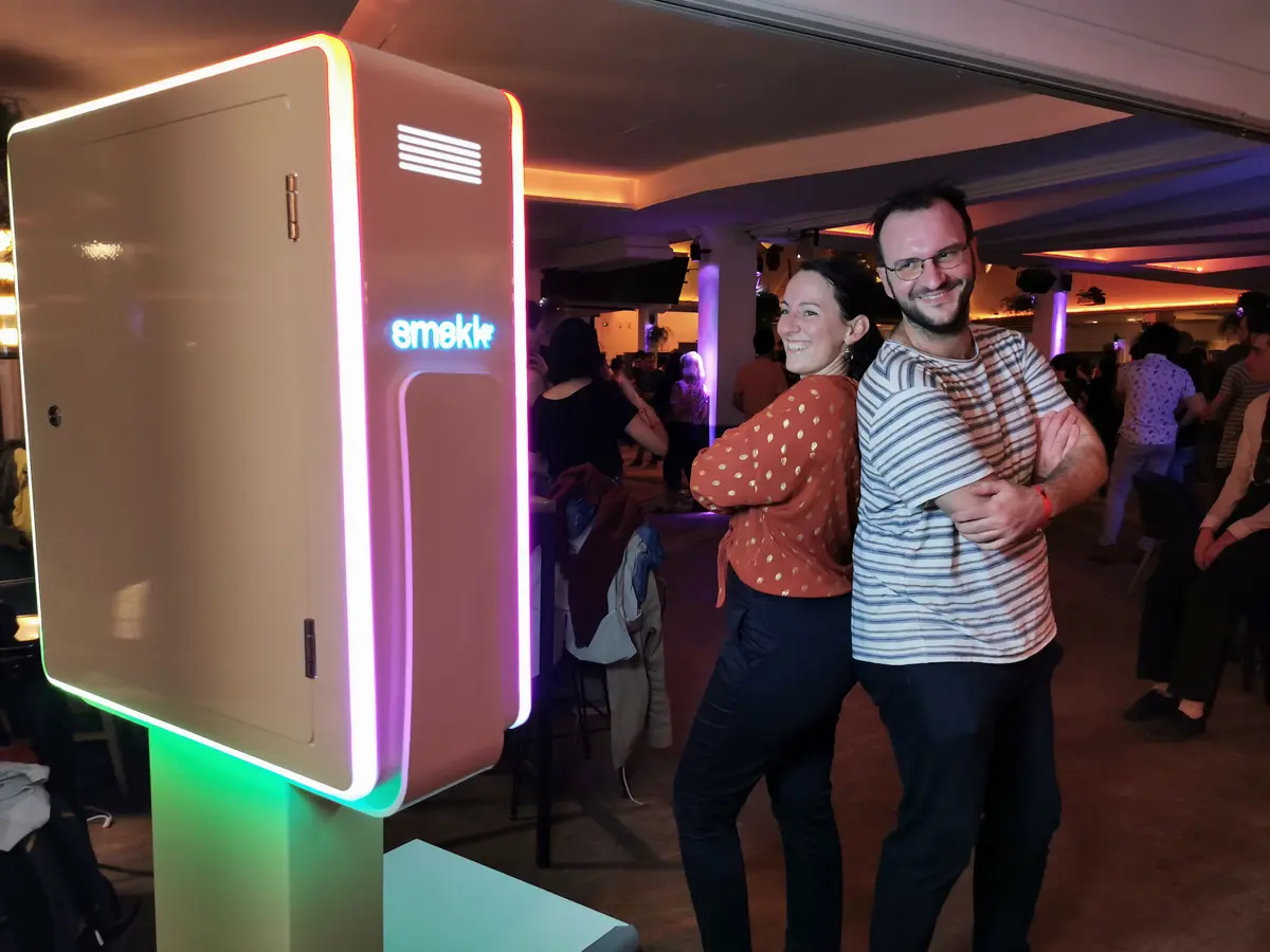 Un couple pose à côté de la borne Smakk éclairée en rose et vert pendant une soirée dansante