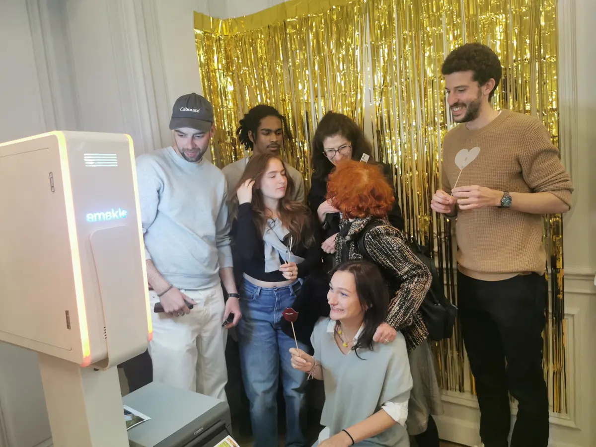 Six invités posent avec des accessoires sur bâtonnet devant un rideau doré, à côté de la borne Smakk