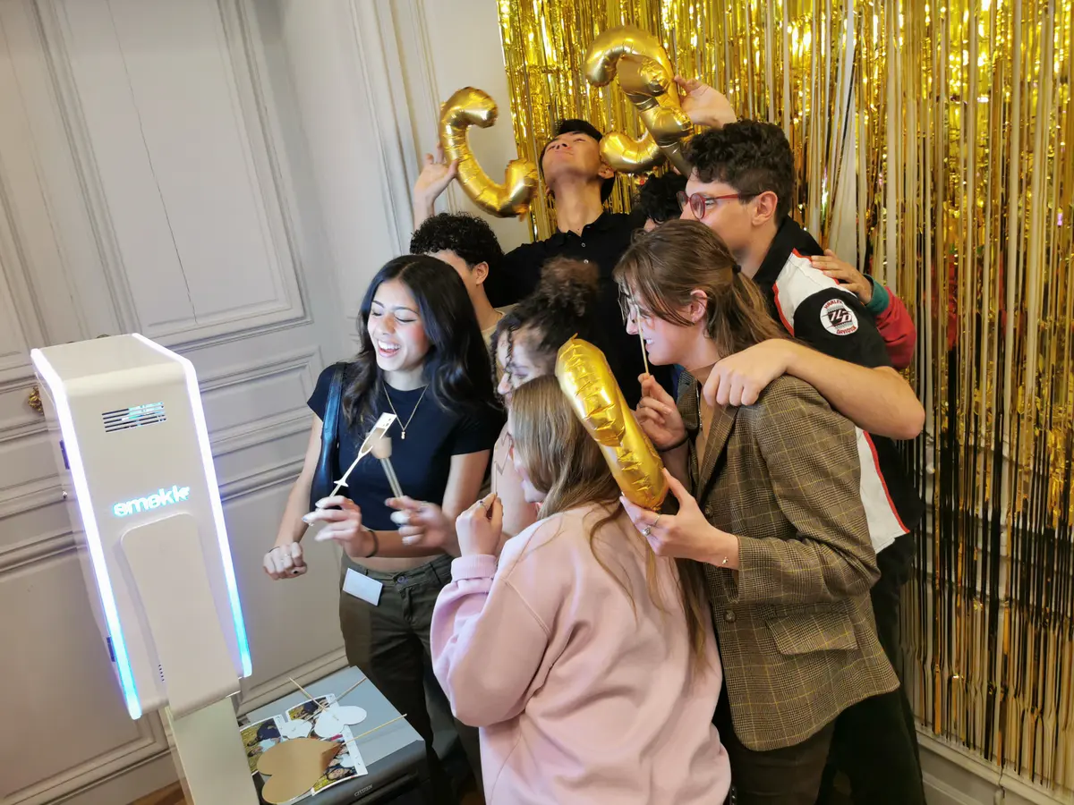 Des invités rient devant un rideau doré avec des ballons en forme de chiffres, la borne Smakk au premier plan